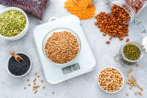 Overhead view of a bowl filled with dried brown lentils placed on a modern kitchen scale. Bowls, bags and containers filled with different legumes are around the kitchen scale and complete the composition.