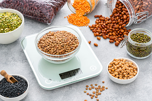 Close up view of a bowl filled with dried brown lentils placed on a modern kitchen scale. Bowls, bags and containers filled with different legumes are around the kitchen scale and complete the composition.