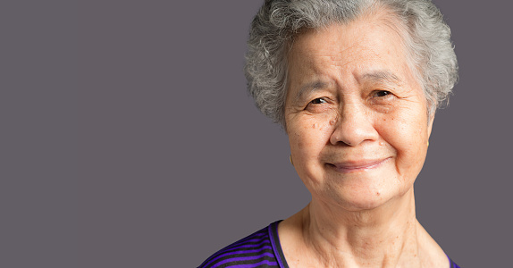 Cheerful an elderly Asian woman with short gray hair looking at the camera with a smile while standing on gray background in the studio. Space for text. Aged people and relaxation concept.