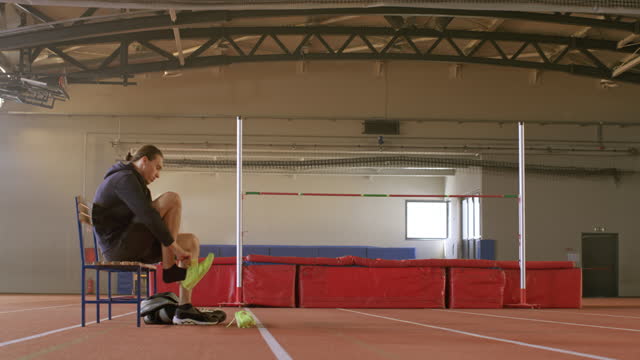 TS Male high jump athlete sitting on the bench in the gym and changing his shoes for practice
