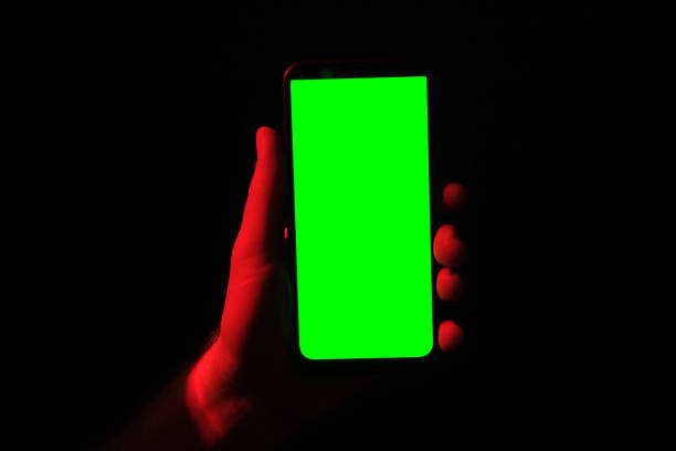 istock Mobile phone in young man hand. Use smartphone with green screen on red blue neon lighting, dark background. Male showing black phone chroma key display. Close up. Hand lifts device up holds it still 1491484521
