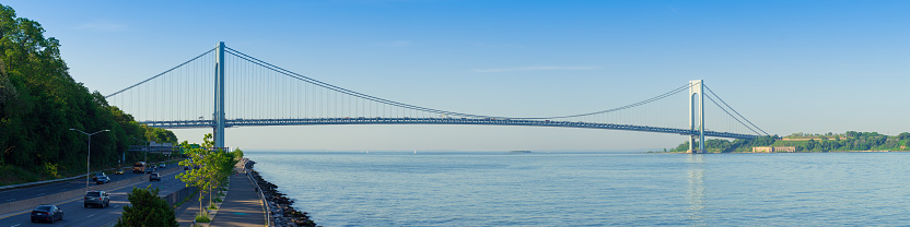 High resolution stitched panorama of the Verrazano-Narrows Bridge on a summer morning with Promenade and Sailboats. The bridge connects boroughs of Brooklyn and Staten Island in New York City. The bridge was built in 1964 and is the largest suspension bridge in the USA. Historic Fort Wadsworth is under the bridge. The photo was taken from the Shore promenade in Bay Ridge Brooklyn. Canon EOS 6D full frame censor camera. Canon EF 85mm F/1.8 lens. 4:1 Image Aspect Ratio. This image was downsized to 50MP. Original image resolution was 66MP or 16251 x 4063 px.