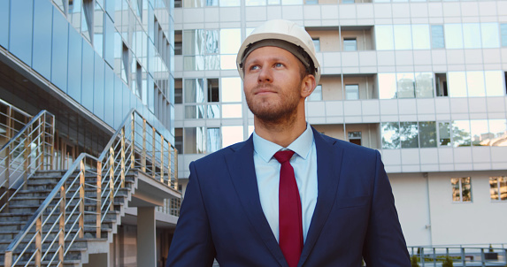 Confident construction manager wearing hardhat walking outside modern building. Young businessman in helmet and suit inspecting construction site outdoors