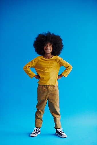 A young black boy with an afro is pictured in a full-length portrait with his hands on his hips, a smile on his face, and a yellow shirt. He is standing in front of a blue background.
