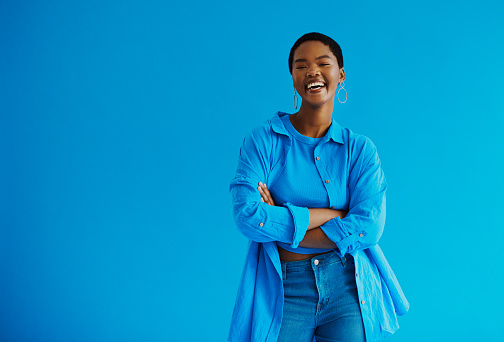 Young black woman standing with her arms crossed, laughing looking at the camera wearing casual clothing, shot against a blue background with copy space stock photo
