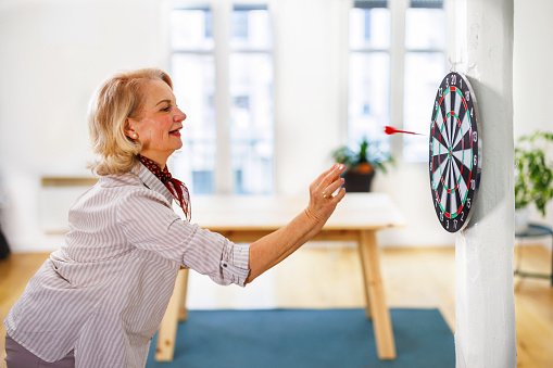 An elderly woman is playing darts in a modern apartment of the house. An older woman is throwing a dart at the dartboard.