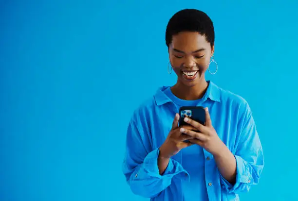 Photo of Young black woman standing and looking at her mobile phone, whilst smiling as she responds back to a text message wearing casual clothing with copy space, stock photo