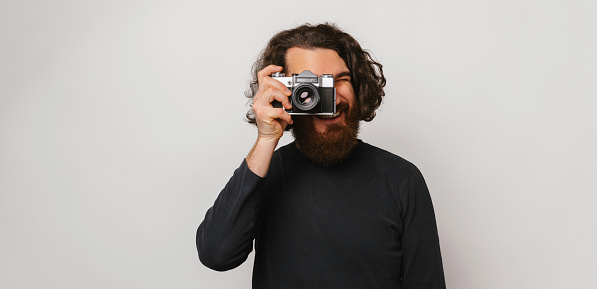 Banner size shot of a long hair bearded man looking through a photo camera over grey background.