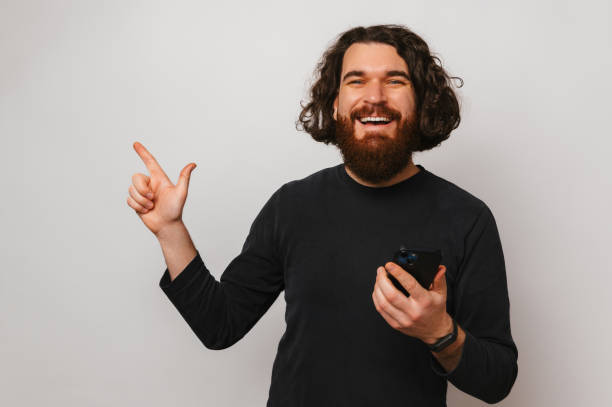 istock Happy bearded man holding his phone is pointing aside at grey copy space. 1491499950