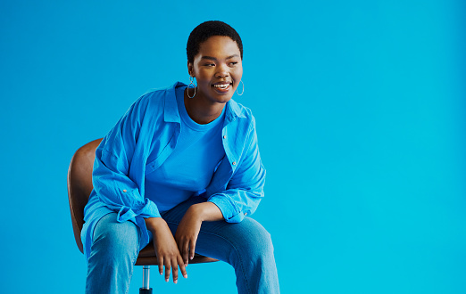 Young black woman sitting on a chair whilst leaning to the side, looking away from the camera whilst smiling with copy space, stock photo