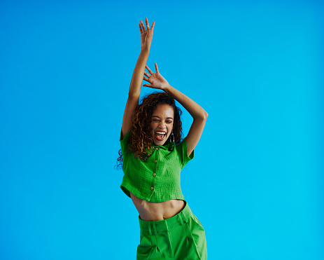 Young multiracial woman dancing moving side to side wearing a green shirt and green pants with her eyes closed and  arms raised, shot against a blue background with copy space, stock photo
