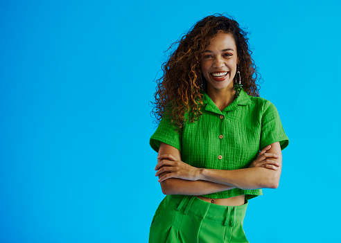 Beautiful young multiracial woman with long curly hair smiling and laughing with her mouth open looking at the camera whilst standing with her arms crossed wearing a green shirt and green pants with copy space, stock photo