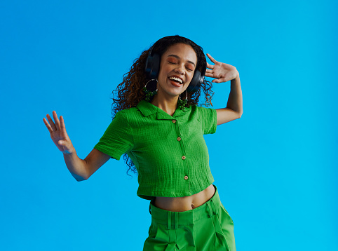 Beautiful dancing multiracial young woman, having fun whilst listening to music wearing a green shirt and green pants with her eyes closed shot against a blue background with copy space, stock photo