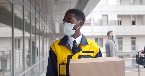 Afro-american courier in safety mask delivery package in modern office building. african-american delivery guy wearing protective mask carrying parcel box walking outside business center