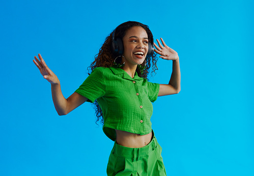 Beautiful dancing multiracial young woman, having fun whilst listening to music wearing a green shirt and green pants whilst looking away from the camera shot against a blue background with copy space, stock photo
