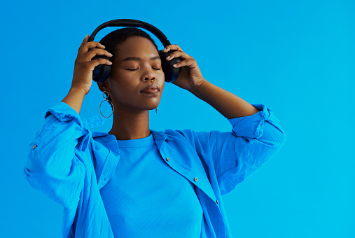 Young black woman standing putting her headphones on with her eyes closed whilst wearing casual clothing shot against a blue background with cop space, stock photo