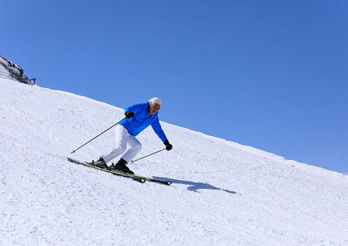 Vital senior, gray-haired men snow skier skiing, enjoying on sunny ski resorts. Skiing carving at high speed against blue sky.