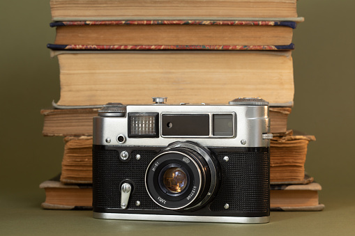 An old analog camera and old yellowed books stacked behind the camera. Items on a green olive background.