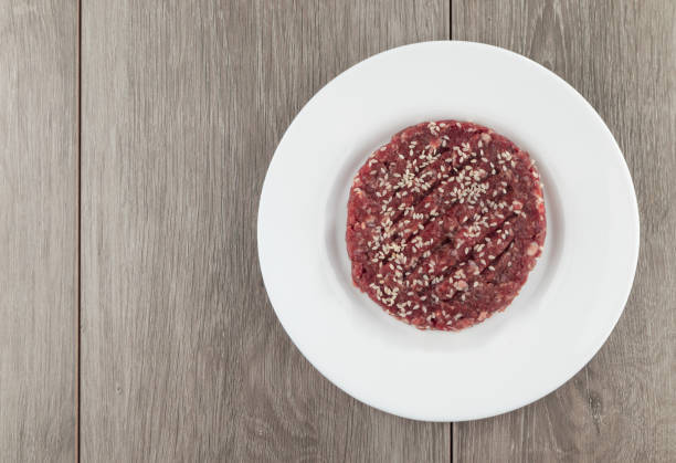 istock Raw hamburger steak with sesame seeds on white plate over wooden background. 1491520403