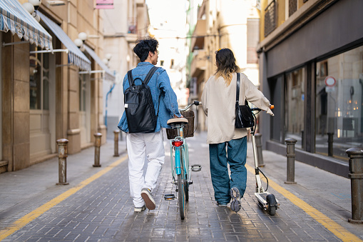 Rear-view of an unrecognizable Chinese woman and man, commuting with bicycle and electric push scooter through Alicante