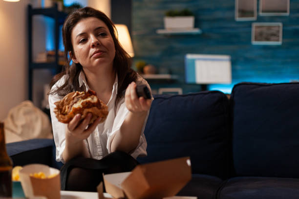 istock Caucasian female holding tasty burger in hands changing channels using remote watching comedy series 1491541017