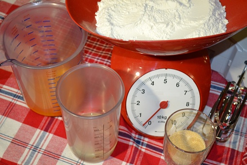 Front top view of different kitchen cooking tools placed on red checkered white tablecloth to start cake cooking: kitchen scale with wheat flour, mixer, transparent plastic cups with fresh eggs and brown sugar.