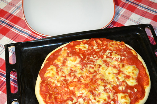 Margherita pizza on a black baking sheet and a large white pizza plate near it. Red checked tablecloth as a background.