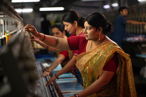 Indian female coworkers dressed in saris operating equipment producing spools in textile factory and representing women empowerment