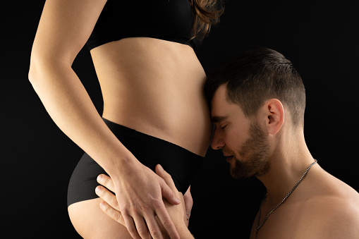 Description: View of pregnant woman's belly with happy father leaning on it with his forehead. Pregnancy first trimester - week 18. Side view. Black background. Bright shot.