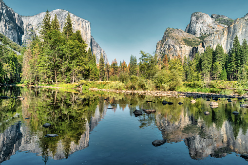 Landscape image of El Capitan and Half Dome in Yosemite National Park with the rocky shore of the Merced River in the foreground, with a vibrant blue cloudy sky.