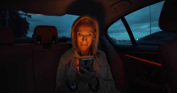 Mature businesswoman sitting on backseat of car and text message. Senior elegant female taking taxi in evening and using smartphone
