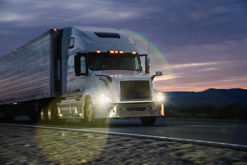 A lone semi-truck traversed the vast expanse of the Nevada Desert, its headlights cutting through the twilight. Against the backdrop of the setting sun, the mountains stood majestic, casting long shadows across the barren landscape. Dust swirled in the wake of the truck, dancing in the fading light. The driver's silhouette was framed by the enormous wheels as the engine roared, echoing through the open desert. The scene was one of solitude and determination, as the truck pushed forward, a solitary figure against the rugged beauty of the Nevada Desert, heading towards the horizon where the last rays of sunlight melted into the night.
