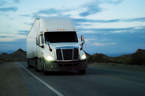 As the sun began its descent, casting a warm golden hue across the Nevada Desert, a semi-truck rumbled through the vast expanse. Dust swirled behind its tires, leaving a trail in its wake. Against the dramatic backdrop of the mountains, their peaks tinged with shades of purple, the truck pressed onward, a lone traveler in this arid landscape. The engine's low rumble harmonized with the wind's gentle howl, creating a symphony of solitude. The driver, eyes fixed on the road ahead, carried the weight of the desert on their shoulders, a stoic figure navigating the ever-changing sands at the edge of daylight.