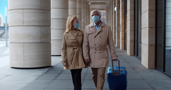 Mature woman and man in medical mask with suitcase walking outdoors. Portrait of senior couple tourist in safety mask with luggage walk outside