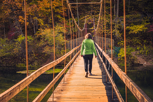Mature woman in green sweater walking along suspension bridge in autumn in Midwestern park; colorful autumn landscape in background