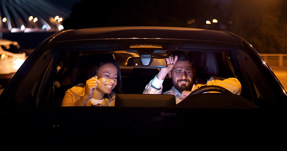 Happy diverse couple sit in car, listen to music and dancing. View through windscreen of woman and man on date in car having fun and dancing late at night