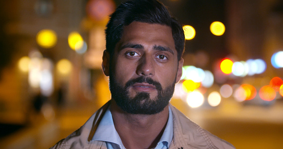 Bokeh shot of handsome indian man looking at camera in city at night. Close up portrait of young indian businessman posing at camera on street against blur lights background