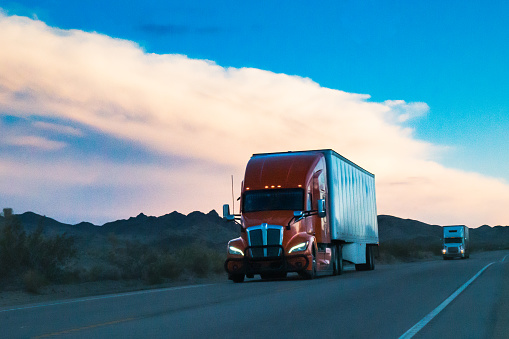 Against the backdrop of a mesmerizing sunset, a colossal semi truck rumbled through the vast expanse of the Nevada Desert. Dust billowed in its wake as it carved its path through the arid landscape. The majestic mountains stood stoically in the distance, their peaks tinged with hues of orange and purple. The truck pressed forward, its metallic frame reflecting the last rays of the setting sun. A solitary figure, the trucker, guided the massive vehicle along the open road, a nomad traversing this desert realm. The scene embodied a sense of freedom and adventure, an ode to the untamed beauty of the American West.