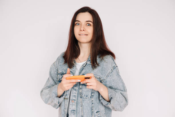 portrait of a young woman in a casual denim jacket with a smartphone playing a game, isolated against a gray background, conveying indecision and insecurity - bolero jacket imagens e fotografias de stock
