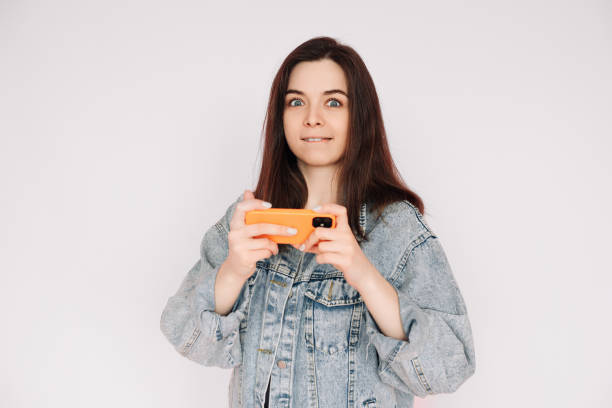 indecisive and insecure: young woman in denim jacket playing smartphone game, isolated on gray background. emphasizing uncertainty and doubt - bolero jacket imagens e fotografias de stock