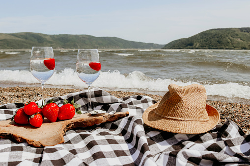 Beautiful sea landscape. Decorated with strawberries, wine glasses, fruit, hat and blanket. Summer vibes with sea waves. Sunrays are making idyllic atmosphere.