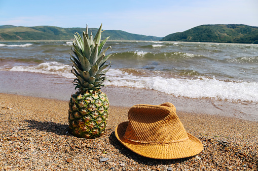 Pineapple in a decoration with a sun hat on the beach. Summer idyllic atmosphere with good vibes. Landed on sand beach with shells and beautiful sea waves in the background.