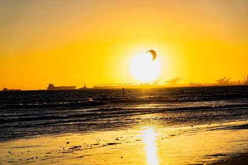 As the sun gracefully descends towards the horizon, casting a golden glow over the port of Los Angeles, a group of kite-surfers takes to the water. Against the backdrop of towering cranes and container ships, their vibrant kites dance with the wind, creating a mesmerizing display of skill and grace.

The kite-surfers, with their boards slicing through the waves, ride the wind with an exhilarating sense of freedom. Their kites, adorned with an array of colors, paint the sky as they soar and twirl with every gust. As the warm hues of the sunset blend with the cool blues of the ocean, the scene becomes a living canvas of contrasting tones.

Cheers and laughter fill the air as the kite-surfers navigate the swells, effortlessly gliding across the water's surface. With each jump and trick, they showcase their mastery of the elements, defying gravity and embracing the thrill of the sport. The sound of the waves crashing against the shoreline harmonizes with the rustling of the kites and the whoosh of the wind, creating a symphony of natural symphony.

Onlookers gather along the shore, their eyes fixed on the spectacle before them. Families, couples, and friends marvel at the spectacle, pointing and capturing the magical scene with their cameras. The atmosphere is electric, charged with a shared sense of awe and admiration.

As the sun finally dips below the horizon, casting a soft, warm glow across the water, the kite-surfers gradually make their way back to the beach. They gather their gear, their faces still flushed with excitement, and exchange tales of their daring maneuvers.