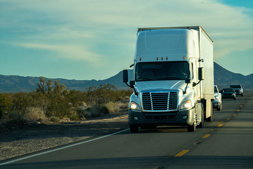 A lone semi truck rumbled through the vast expanse of the Nevada Desert, its powerful engine echoing against the arid landscape. As the sun descended towards the horizon, casting a warm golden glow, the majestic mountains stood tall in the distance, their peaks painted with hues of purple and orange. Dust kicked up behind the truck, swirling in the air as if dancing to the rhythm of its tires. The scene exuded a sense of both solitude and determination, as the truck pressed on, carrying its cargo through the untamed beauty of the desert, accompanied by the timeless silhouette of the mountains.