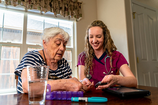 A young female nurse administered a blood sugar test to an elderly woman sitting at a dining room table. The two shared a laugh, making the process seem less daunting and building a bond of trust and companionship.