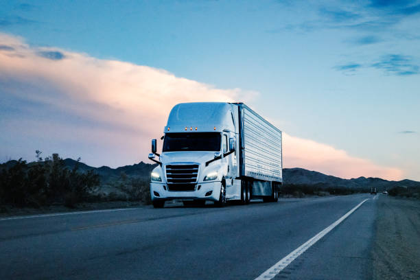 At sunset, a white semi truck rumbled through the vast Nevada Desert. Against the majestic backdrop of mountains, it pressed on, leaving tire tracks on the desert floor. Against the backdrop of a setting sun, a lone semi-truck powered through the vast expanse of the Nevada Desert. Its massive wheels kicked up swirls of dust as it journeyed across the arid landscape. The rugged mountains, silhouetted by the fading light, towered in the distance, their majestic peaks casting long shadows over the desert floor. The truck pressed on, its engine humming a steady rhythm as it carved a path through the deserted terrain. The scene evoked a sense of isolation and resilience, as the truck and mountains stood as testaments to the unforgiving beauty of the Nevada Desert at sunset. semi truck stock pictures, royalty-free photos & images