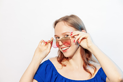 Studio portrait of a happy young white woman with long brown hair in a blue dress, wearing pink sunglasses, against a white background