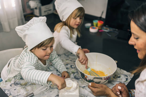 pair of blond brother and sister children baking homemade cupcakes. one of them scoops flour with a spoon and the other one beats eggs. - cake making mixing eggs imagens e fotografias de stock