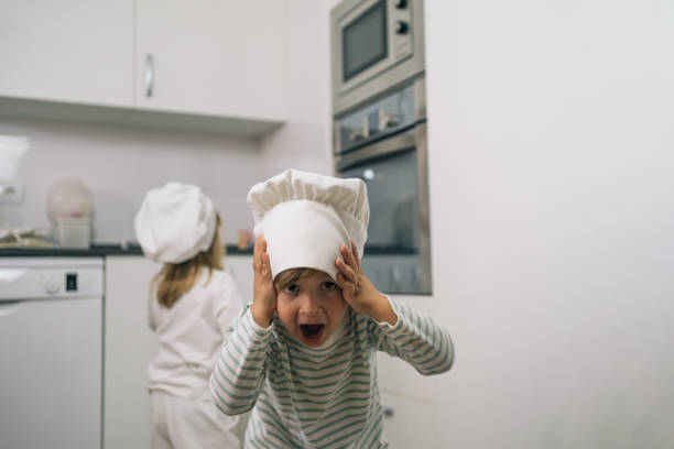 Blond boy in a chef's hat smiling with a surprised face holding his hands to his face while his sister checks the oven.