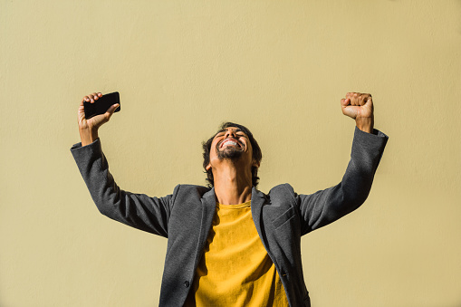 a latin businessman celebrates success with his arms up in the air and looking at the sky with a euphoric smile with his cell phone in his hand making a fist on a yellow background.